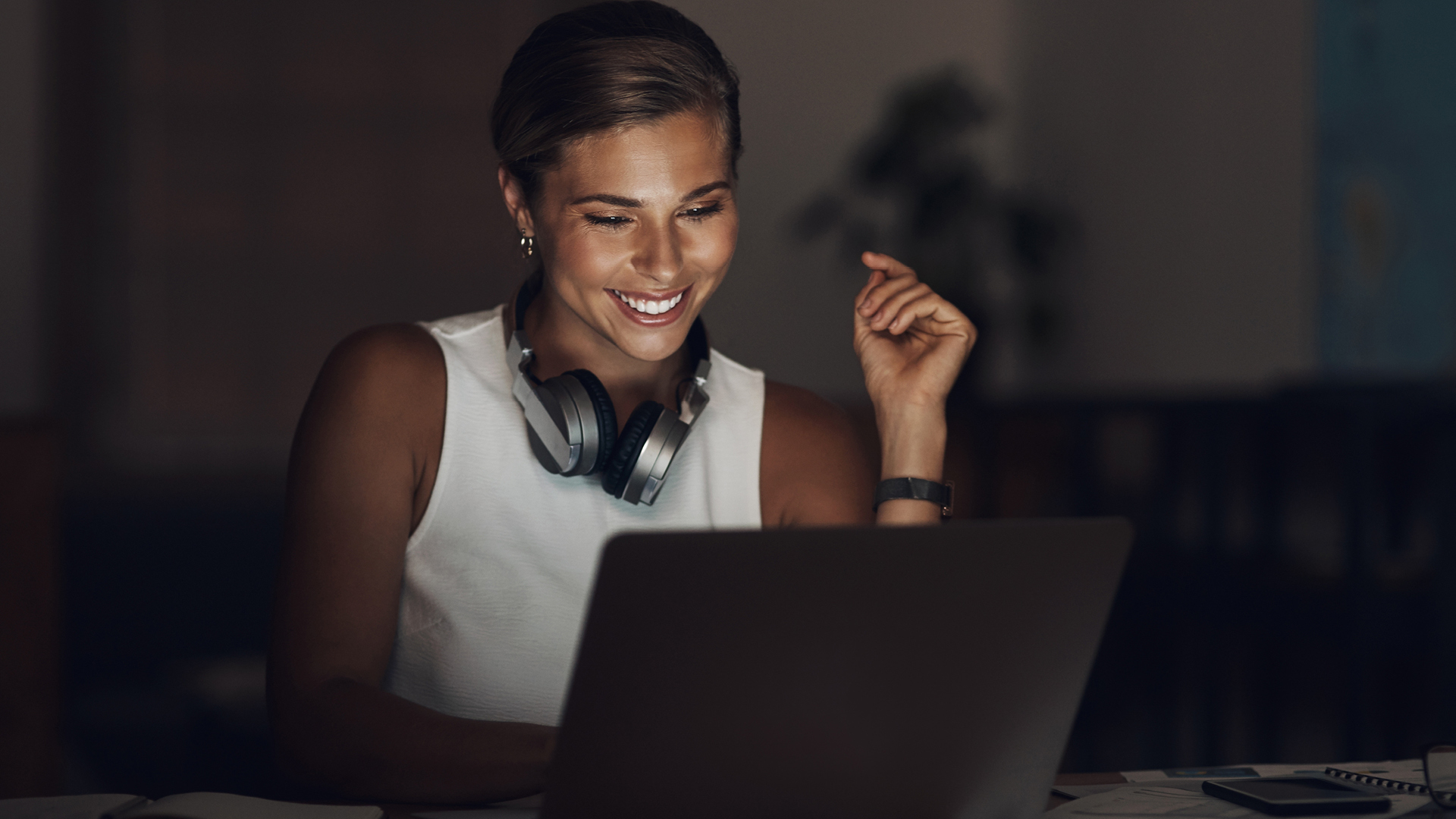 A woman wearing a white sleeveless top and headphones smiles while looking at a laptop screen in a dimly lit room. Her hair is tied back, and her expression appears enthusiastic and engaged.