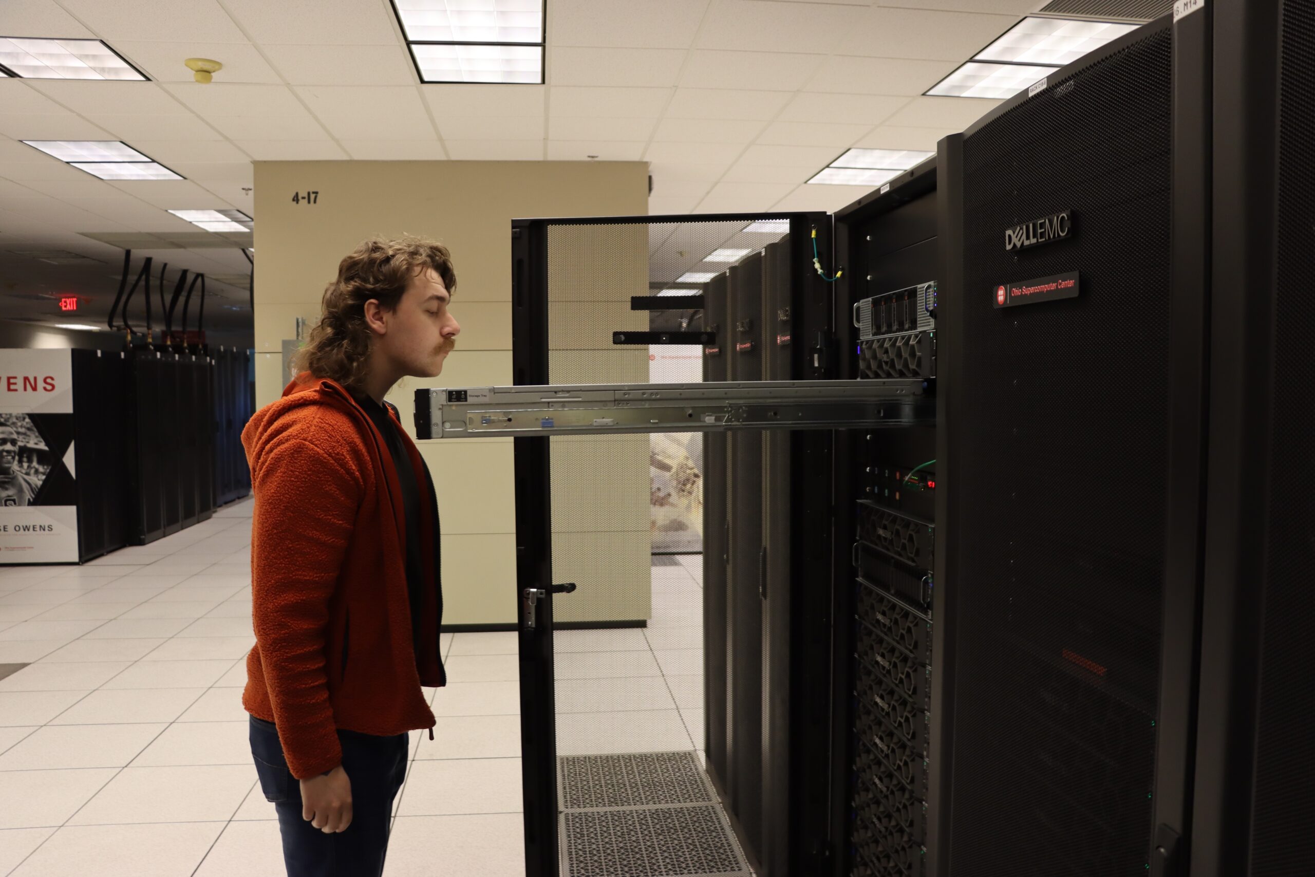 A person in a red hoodie examines a large server rack in a data center. The individual leans in to inspect the equipment closely, with various server units and cables visible. The room is brightly lit with a tiled floor.