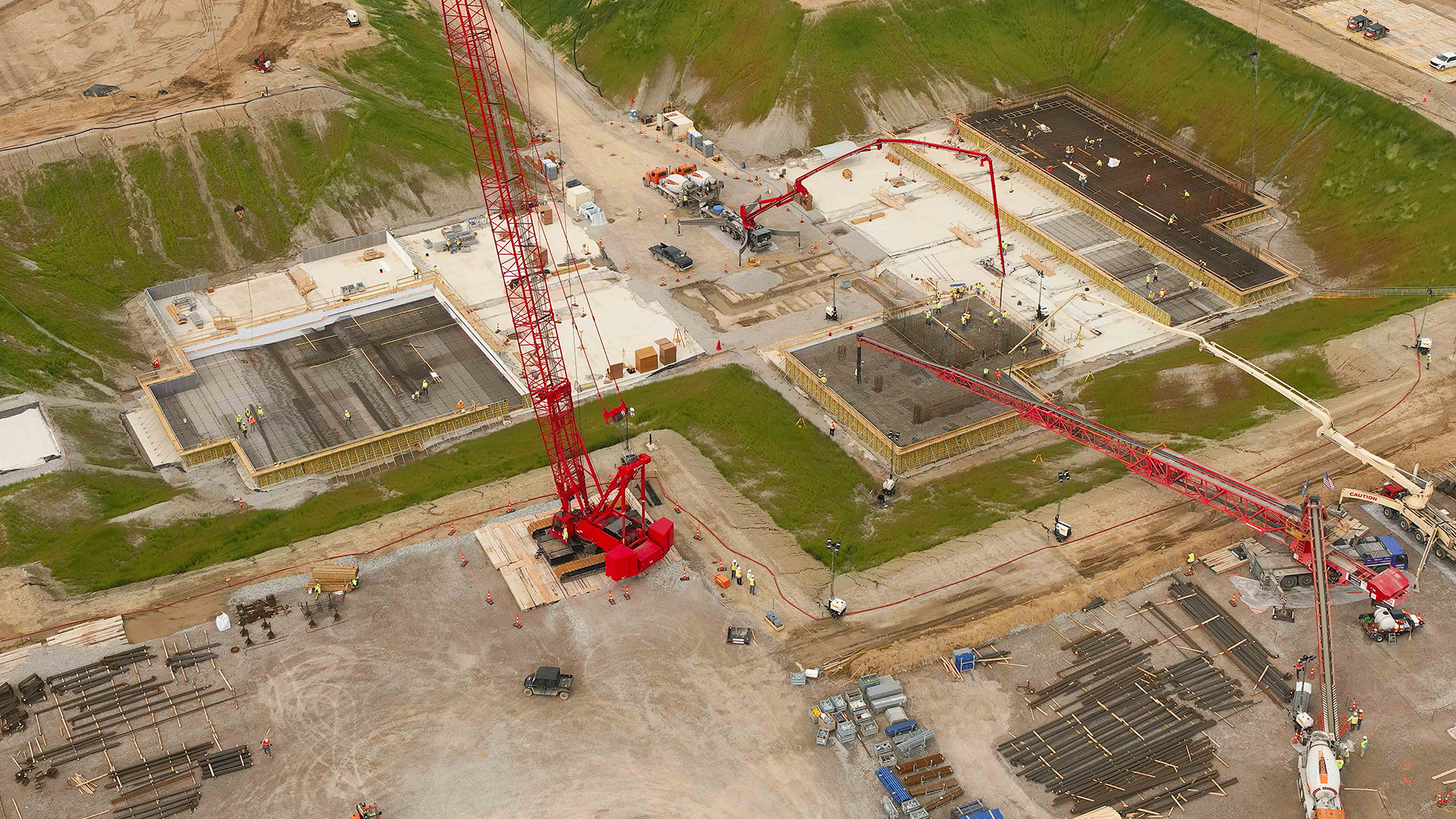 Aerial view of a construction site with a large red crane and multiple concrete foundations. Workers and machinery are visible around the area. Green grassy slopes surround the site. Various materials are scattered throughout the ground.