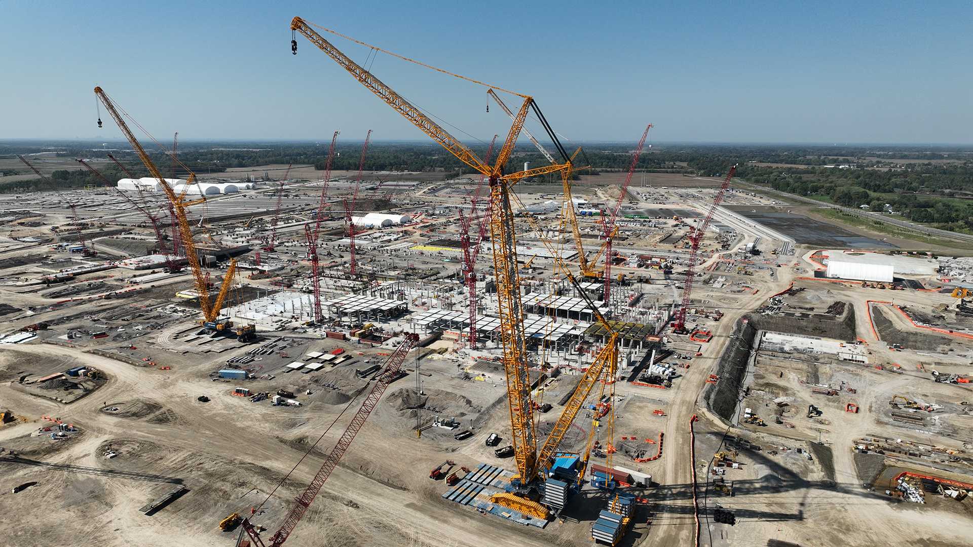 Aerial view of a large construction site with multiple cranes, machinery, and building structures. The landscape is expansive, with clear skies and distant trees on the horizon.