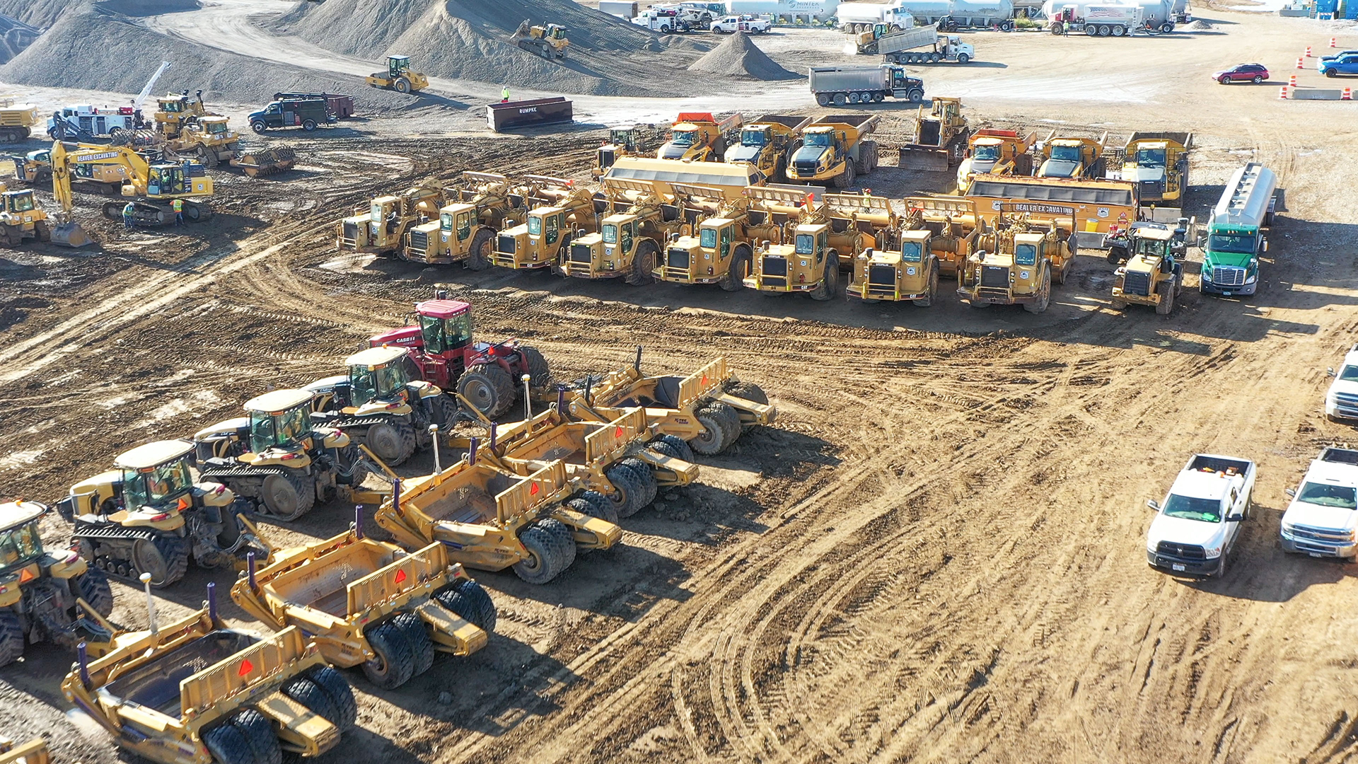 Aerial view of a construction site with multiple yellow heavy machinery vehicles, such as bulldozers and dump trucks, arranged in rows. Several trucks and a red tractor are also visible on the dirt ground. Dirt mounds and equipment are in the background.