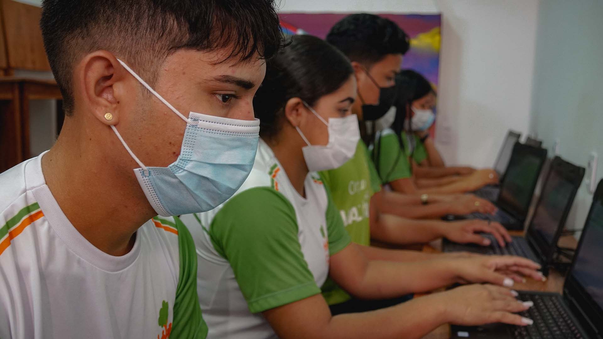 A group of young people wearing masks and green and white shirts are sitting in a row, working on laptops in a classroom setting.