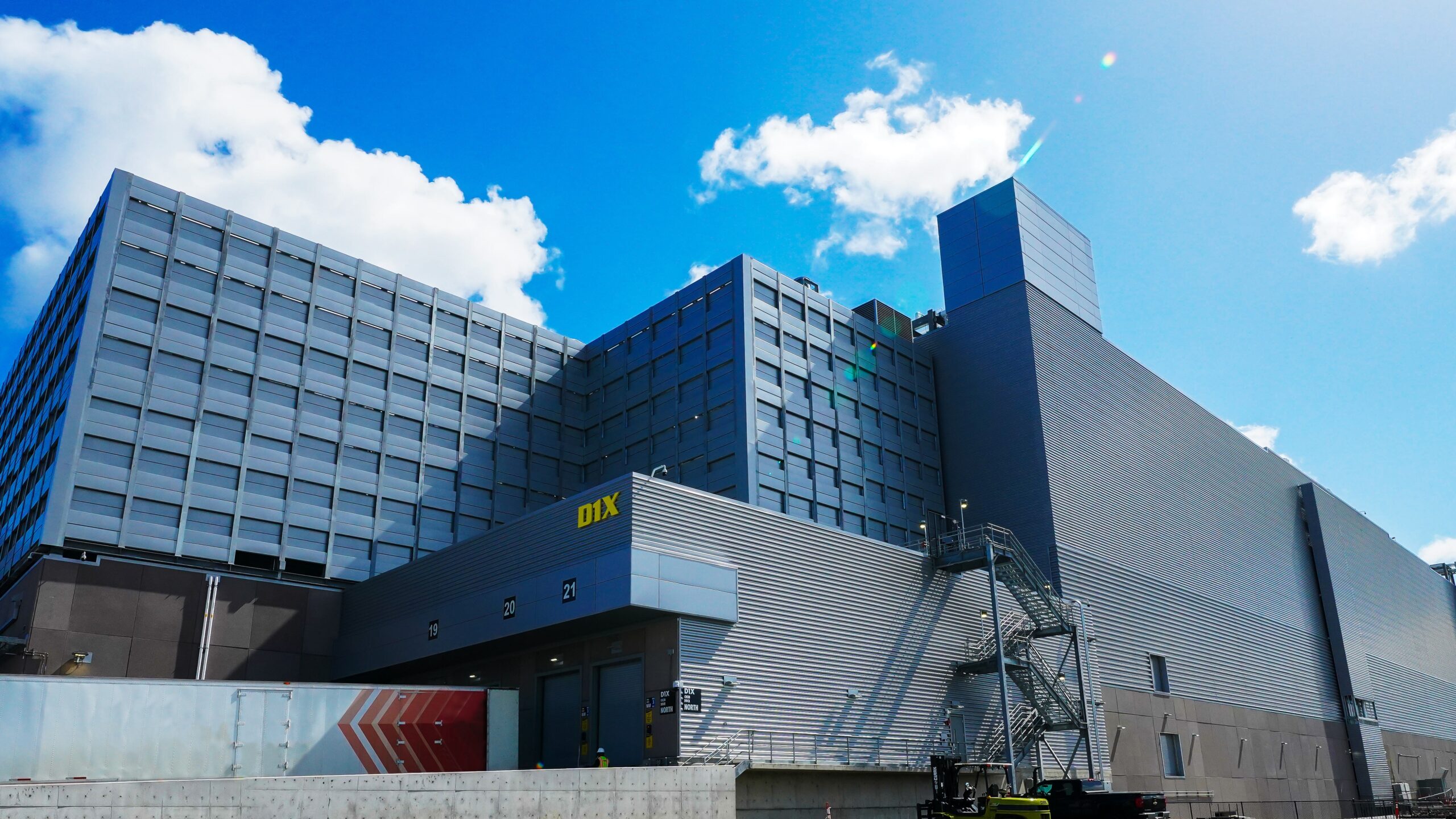 A large industrial building with a gray and blue facade stands under a clear blue sky. The structure features modern architecture, multiple stories, and a visible staircase on the side. A bright sunlight glares from the top right corner.