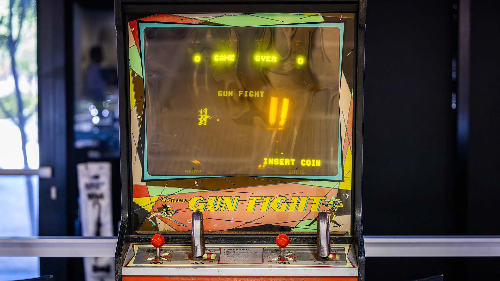 A vintage arcade game machine displaying Game Over and Gun Fight on the screen. The machine has colorful artwork, two red joysticks, and metal buttons, with an Insert Coin prompt.