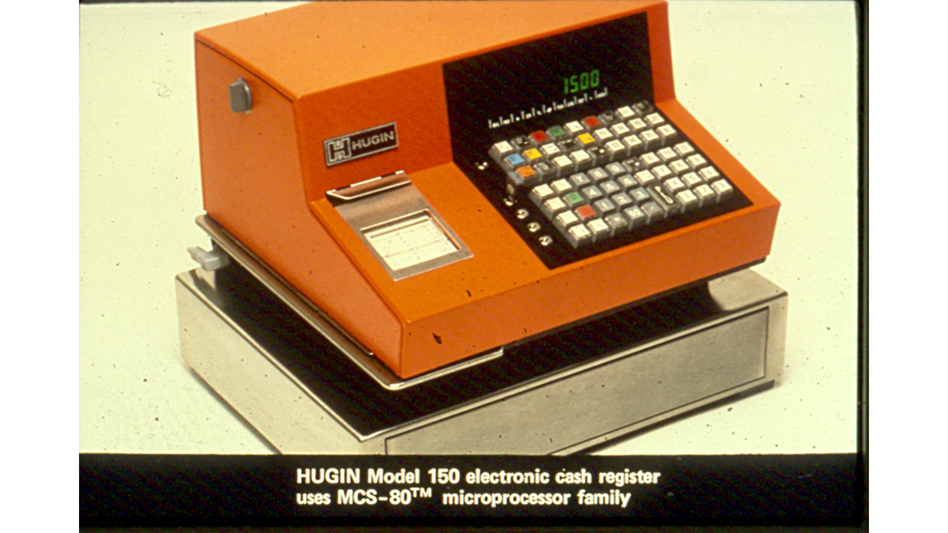 An orange Hugin Model 150 electronic cash register, featuring a digital display, keypad, and receipt printer, set on a metal base. The caption notes it uses the MCS-80 microprocessor family.