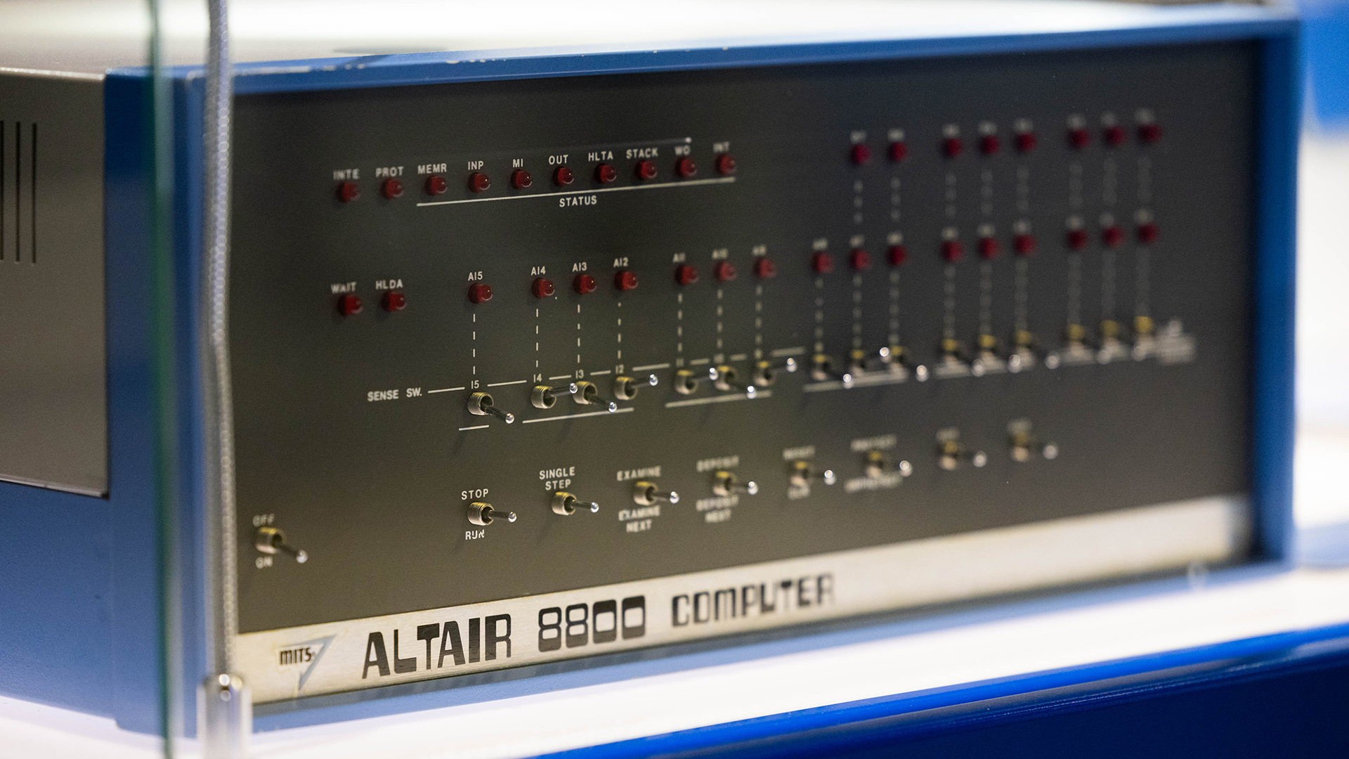 A close-up of a vintage Altair 8800 computer, showcasing its front panel with numerous toggle switches and indicator lights. The metal panel features labels for each switch and light, housed in a blue casing.