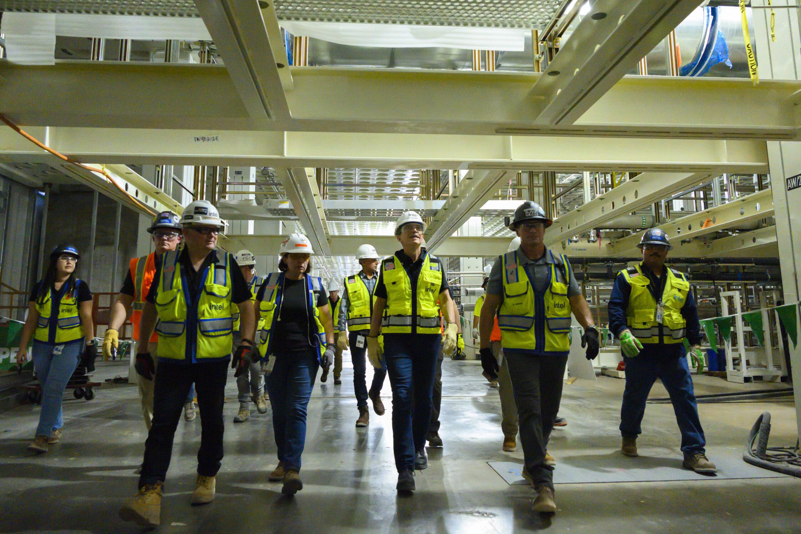 A group of construction workers wearing safety vests and helmets walk through an industrial building. The interior features metal beams and overhead structures. The workers appear focused and are equipped with gloves and protective gear.