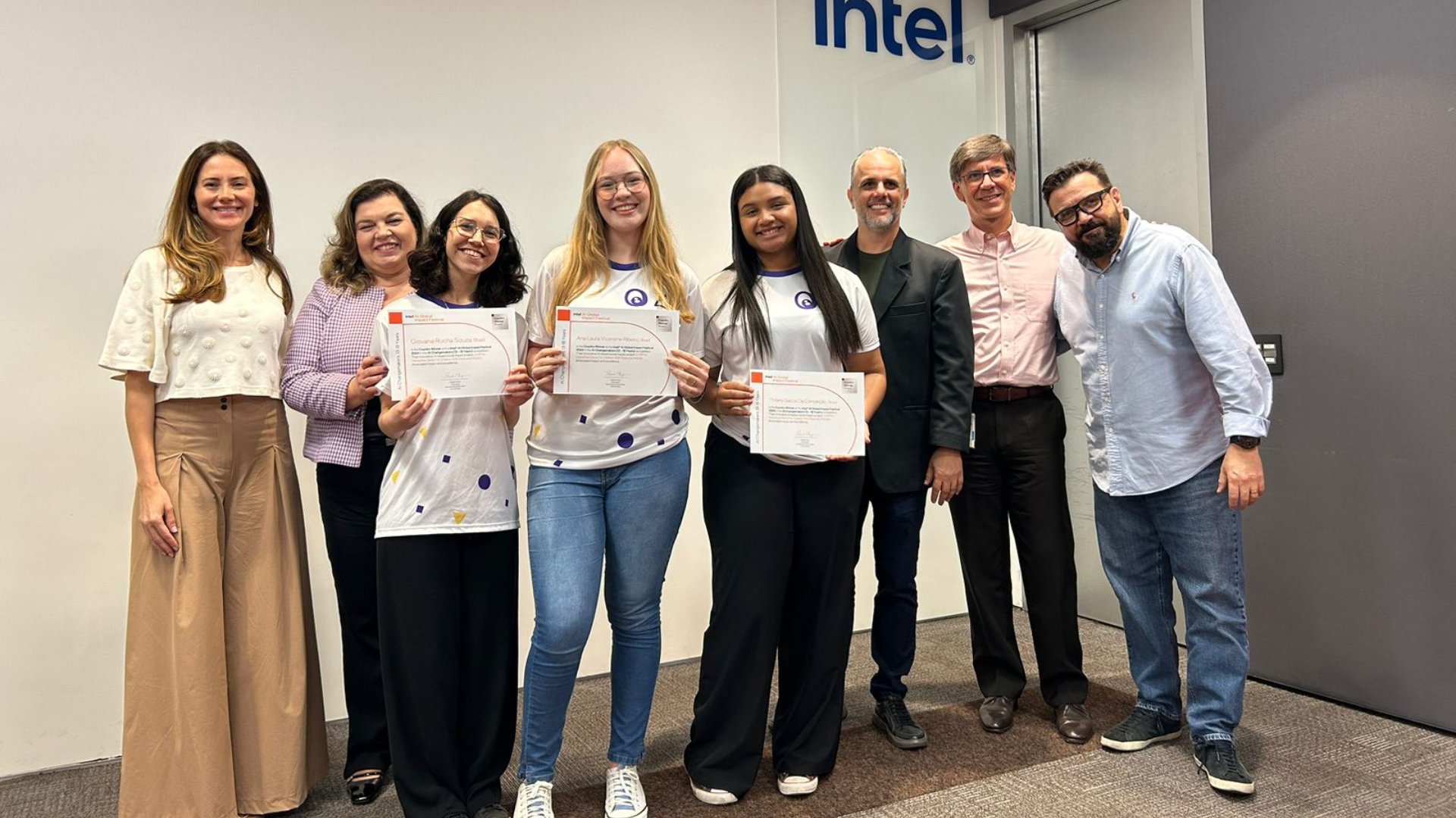 A group of people standing and smiling in an office setting. Three women in the center hold certificates. The group includes both men and women, dressed in business casual attire. A partial Intel logo is visible on the wall behind them.