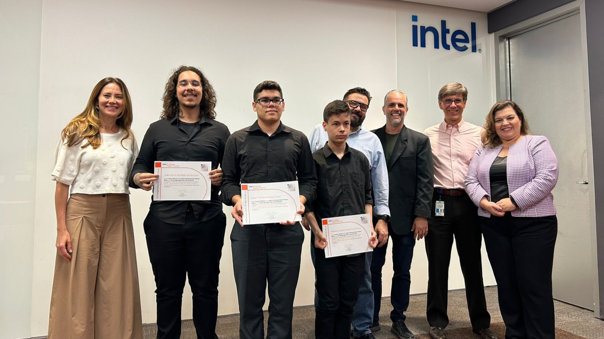 A group of people standing indoors, with three of them holding certificates. The group is positioned in front of a wall with the Intel logo. They appear to be at a formal or celebratory event.