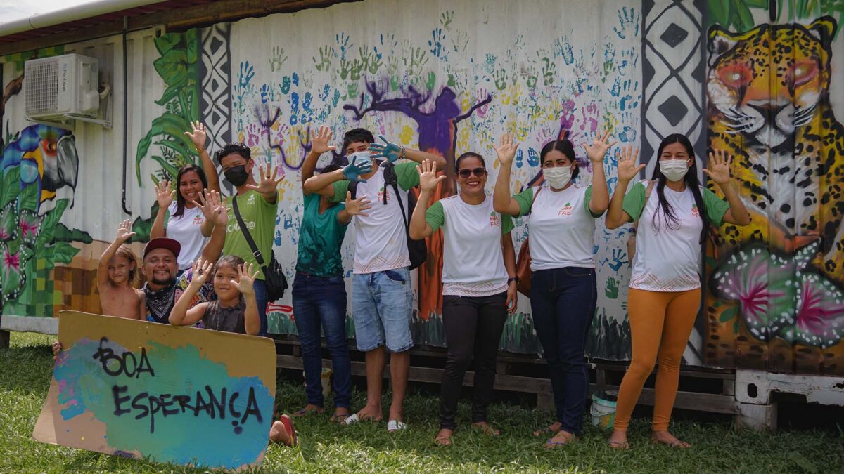 A group of people standing outdoors in front of a colorful mural featuring a jaguar and handprints. Some are wearing masks and all are smiling with their hands raised. A child holds a rustic sign reading Boa Esperança! in vibrant letters.