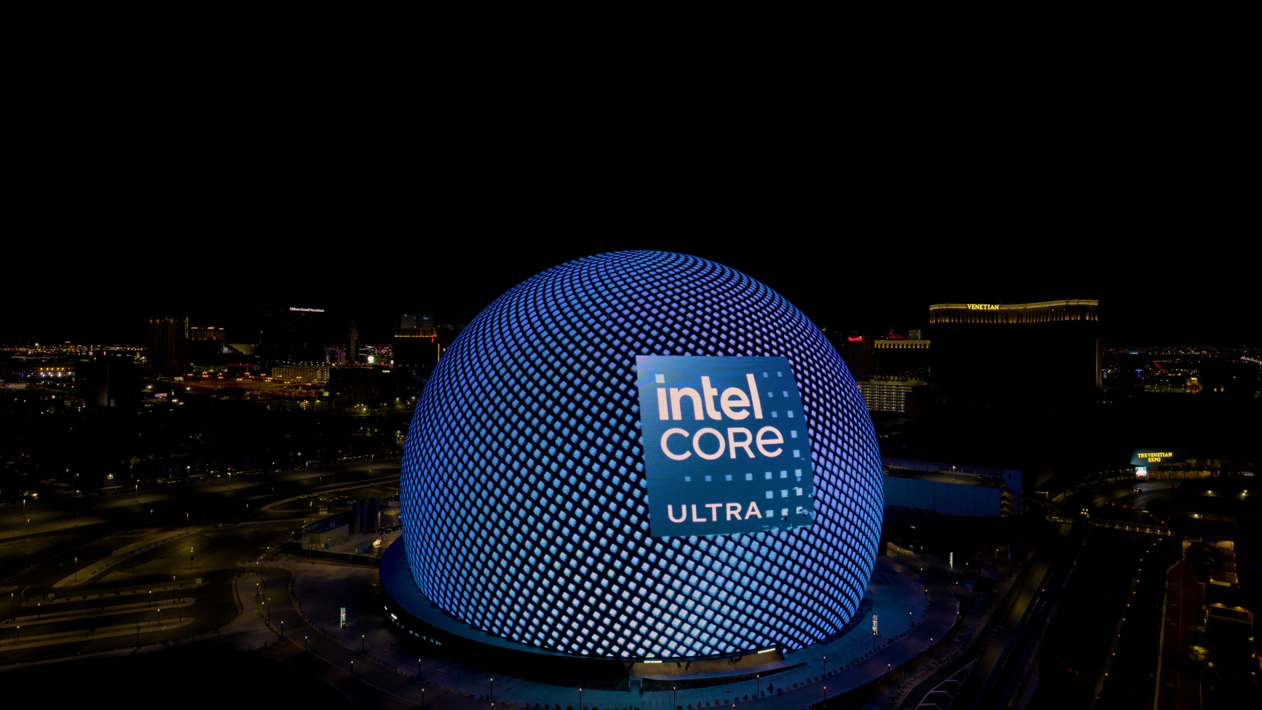 A large spherical building illuminated at night with a blue grid pattern, displaying a prominent Intel Core Ultra logo. The surrounding area is dark with city lights visible in the background.