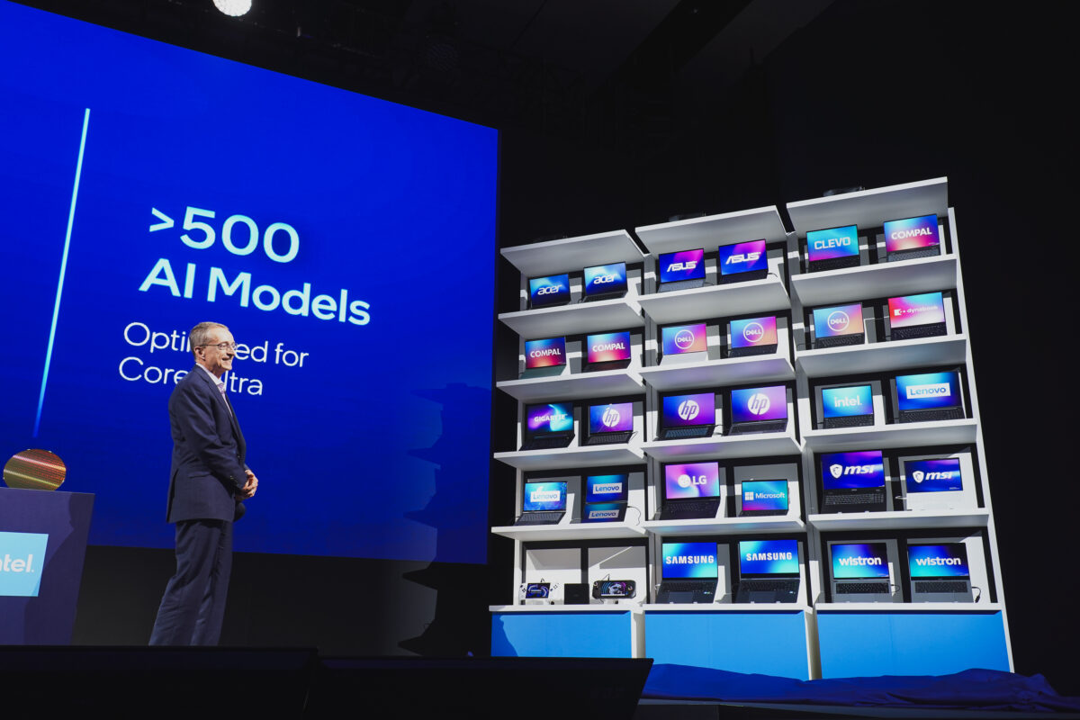 A person stands on stage next to a large screen displaying >500 AI Models Optimized for Core Ultra. Beside them, a wall of monitors shows various tech brand logos like Lenovo, HP, Acer, Samsung, and more, illuminated in vibrant colors.