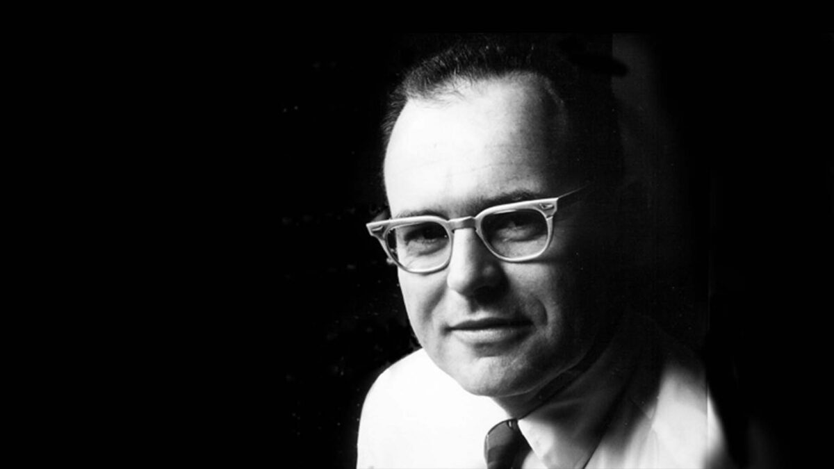 A black and white portrait of a man wearing glasses and a suit, gazing at the camera. The background is dark, emphasizing his face.