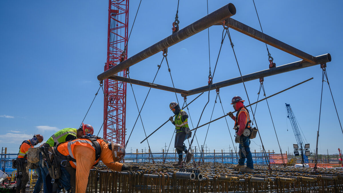 Construction workers wearing helmets and safety gear are assembling a large metal structure at a construction site. A crane supports the structure, and other cranes are visible in the background against a clear blue sky.