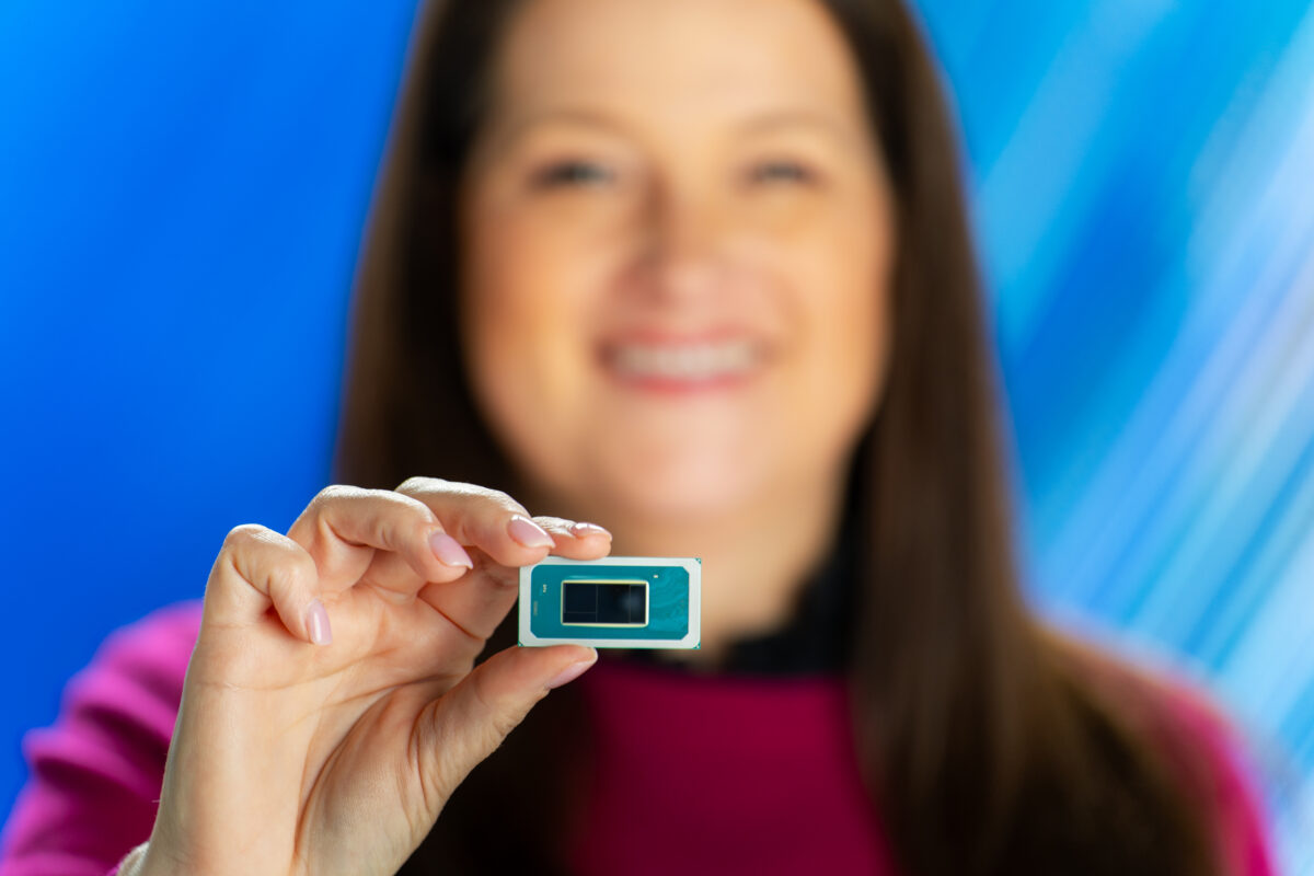 A person with long hair holds a small computer chip between their thumb and index finger. The background is a gradient of blue shades. The person is out of focus, emphasizing the chip.