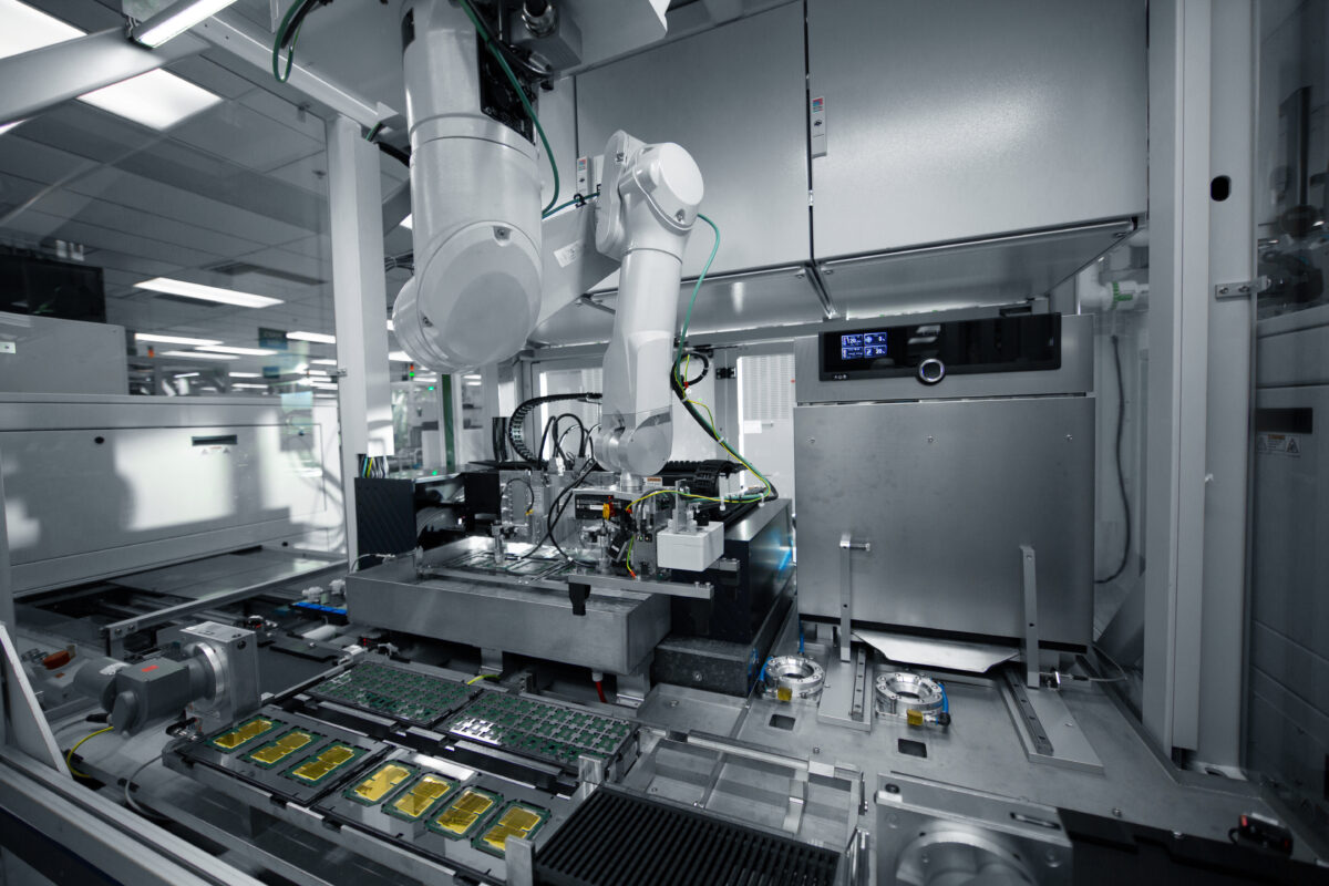 A high-tech laboratory with a robotic arm working on electronic components. The setup includes various machines, wires, and circuit boards under bright overhead lighting, creating a futuristic and technical atmosphere.