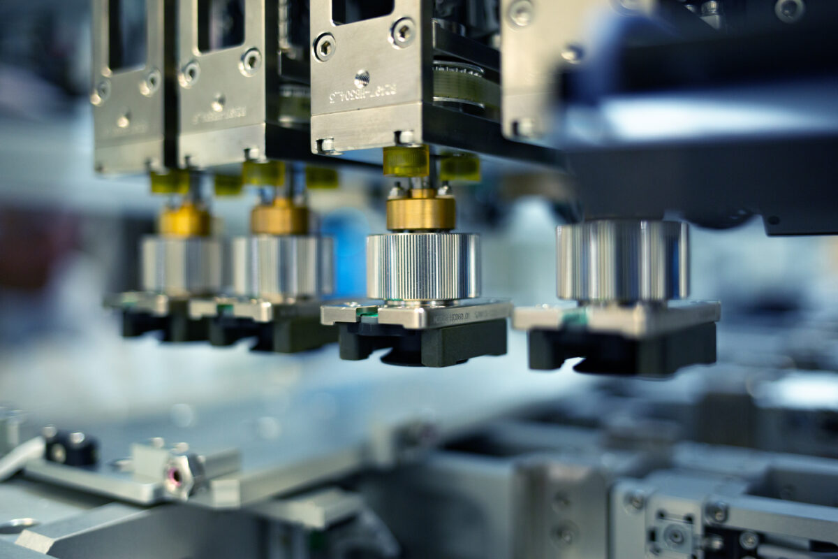 Close-up of automated precision machinery in a factory setting, featuring metallic components and a blurred background. The machine elements appear to be involved in a manufacturing or assembly process.