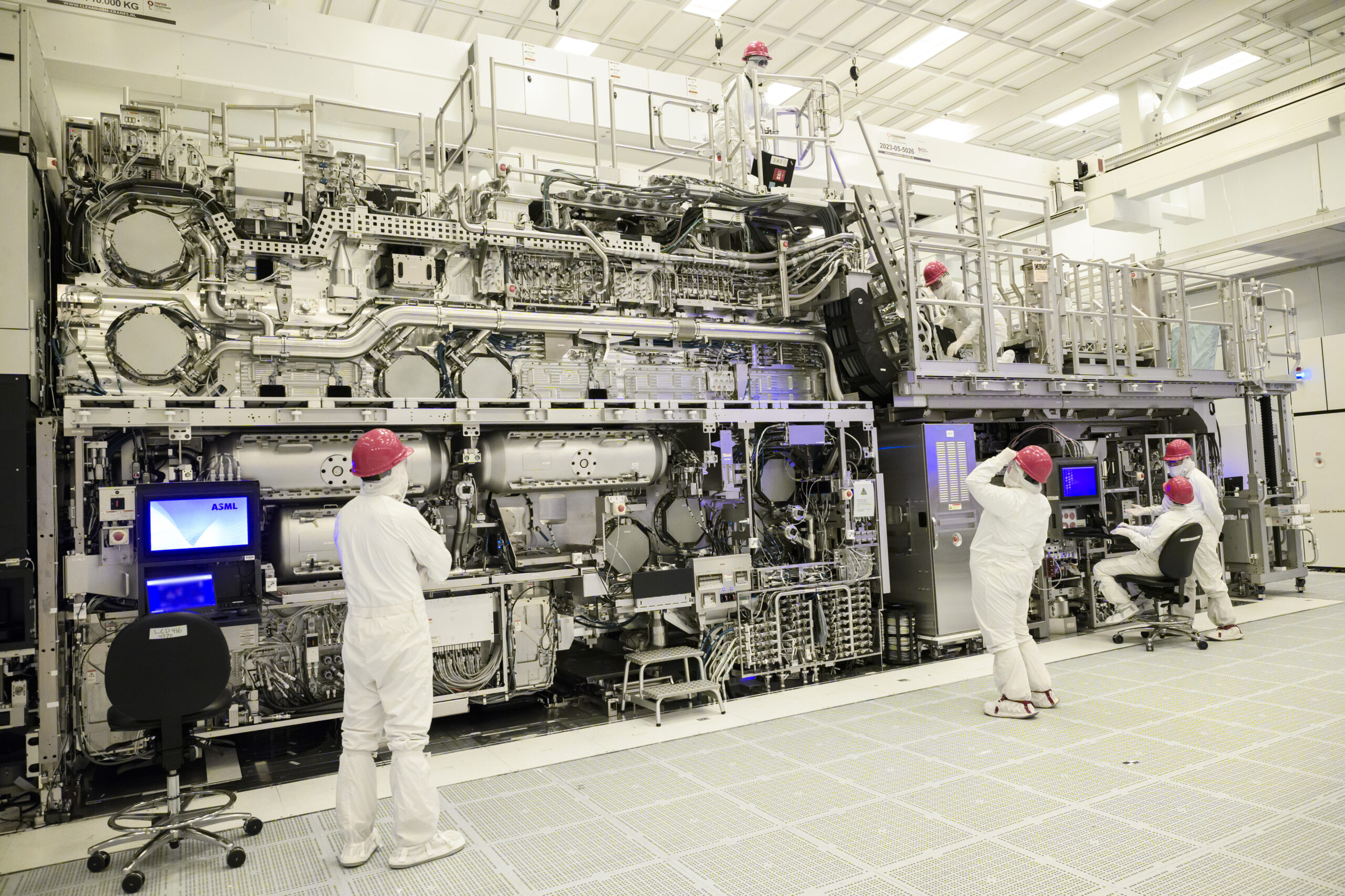 Technicians in white cleanroom suits and red caps work on a large, complex machine with numerous pipes and control panels in an industrial setting. Some use computers, while others inspect the equipment.