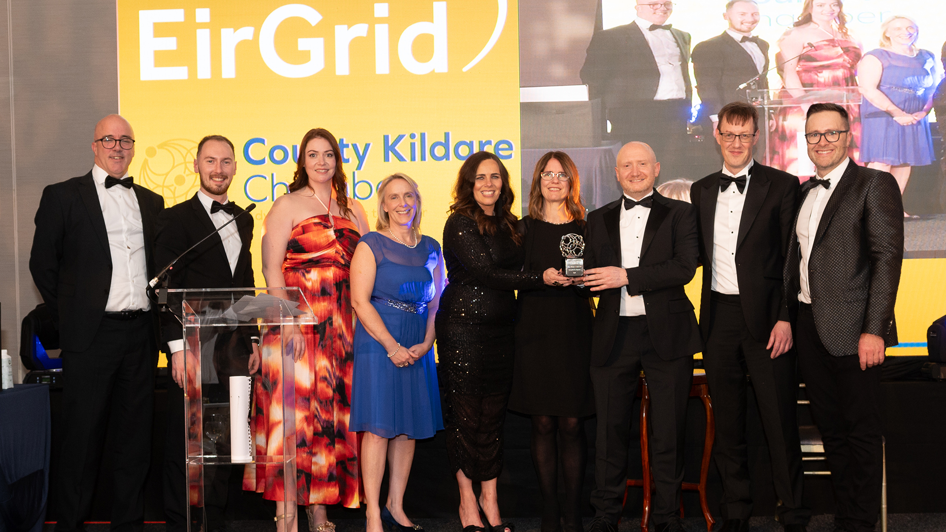 A group of eight people dressed in formal attire pose for a photo at an awards event. They stand in front of a yellow and blue backdrop with the EirGrid logo. One person holds a trophy. A screen in the background displays their image.