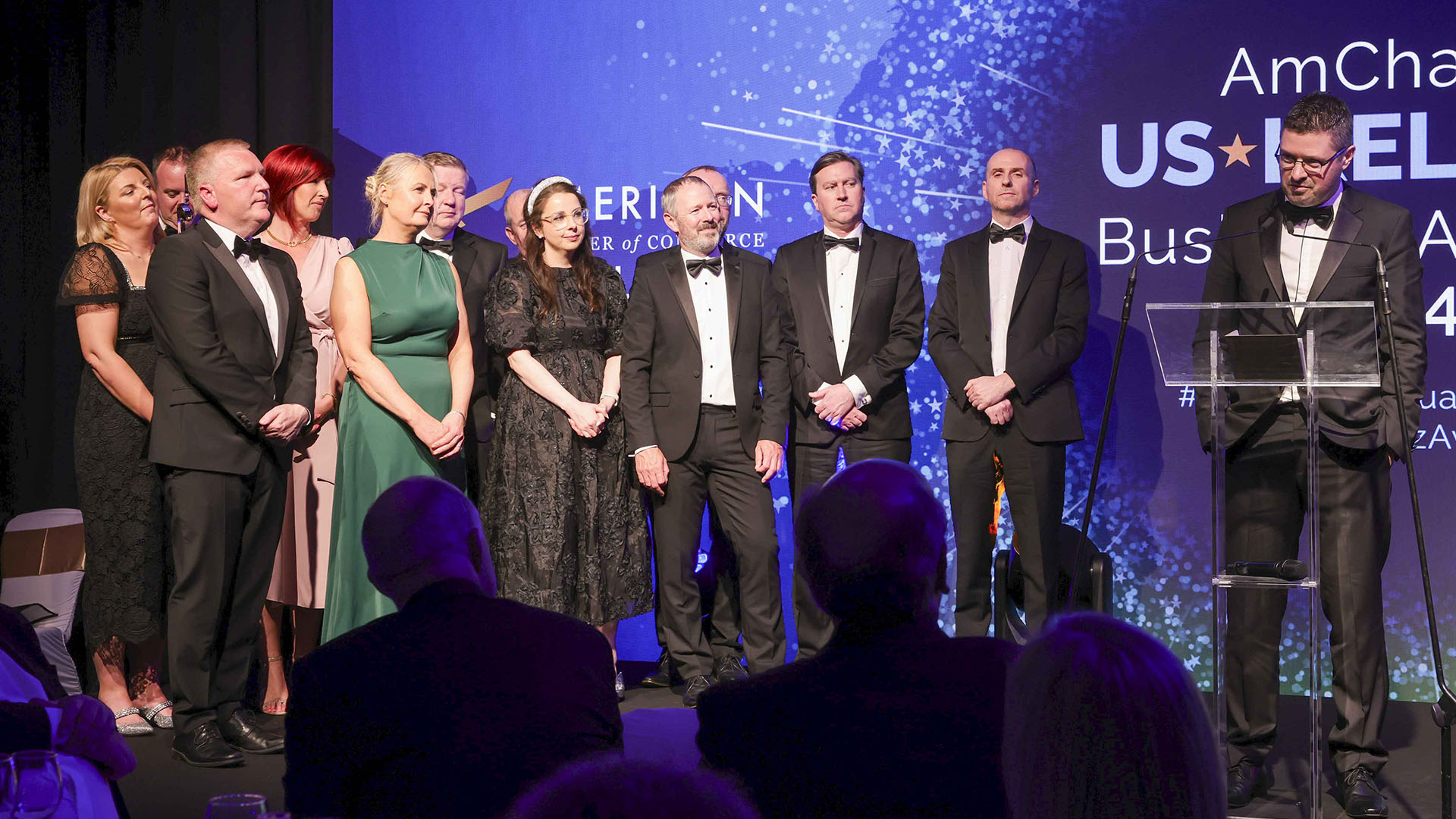 A group of people in formal attire stand on a stage at the AmCham US-EU Business Gala. A man at a podium, wearing glasses and a tuxedo, addresses the audience. A large screen in the background displays event information.