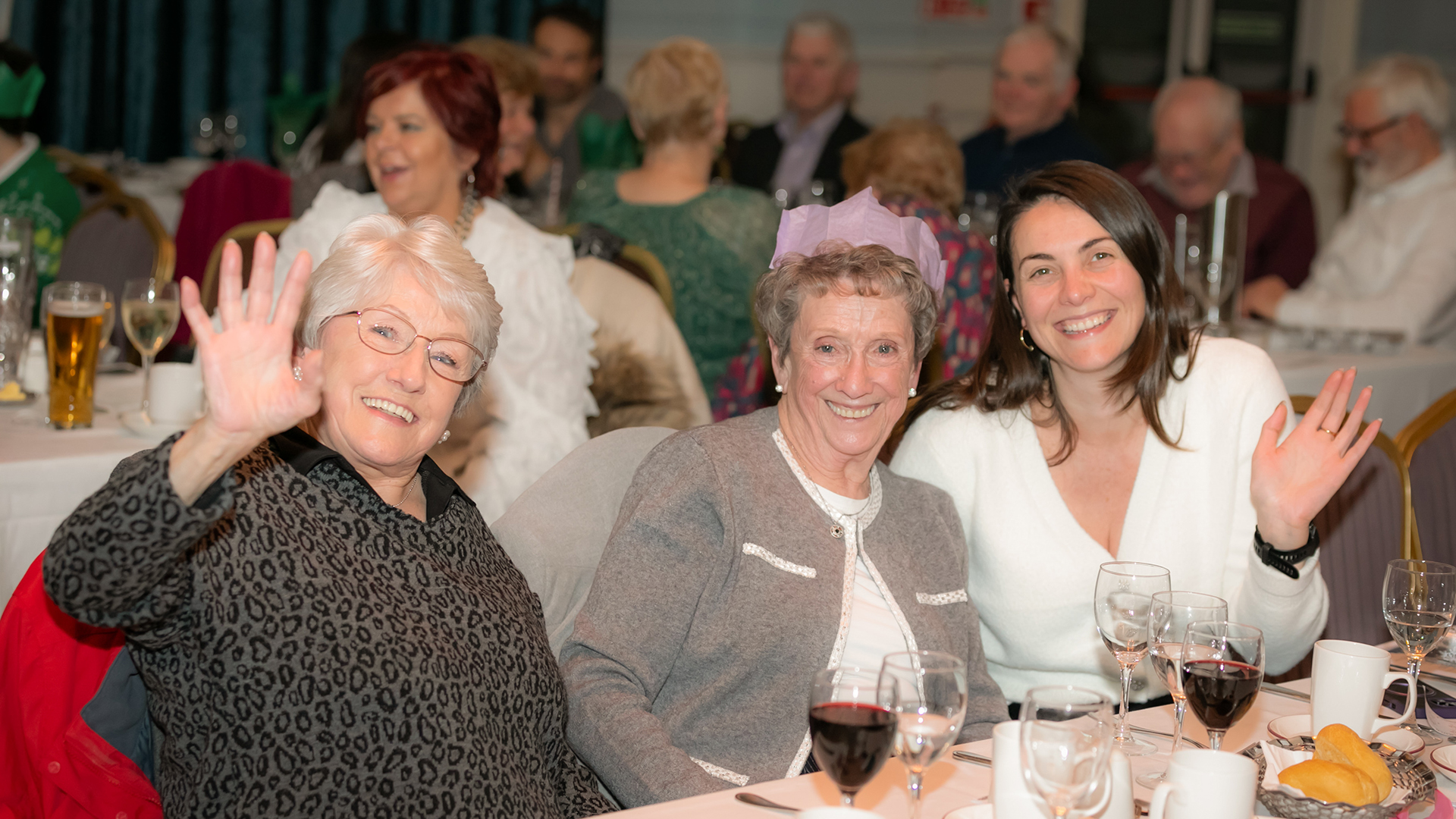 Three women sitting at a table during a festive gathering, smiling and waving at the camera. They are surrounded by glasses of wine and other attendees, some of whom are partially visible in the background, all appearing to enjoy the occasion.