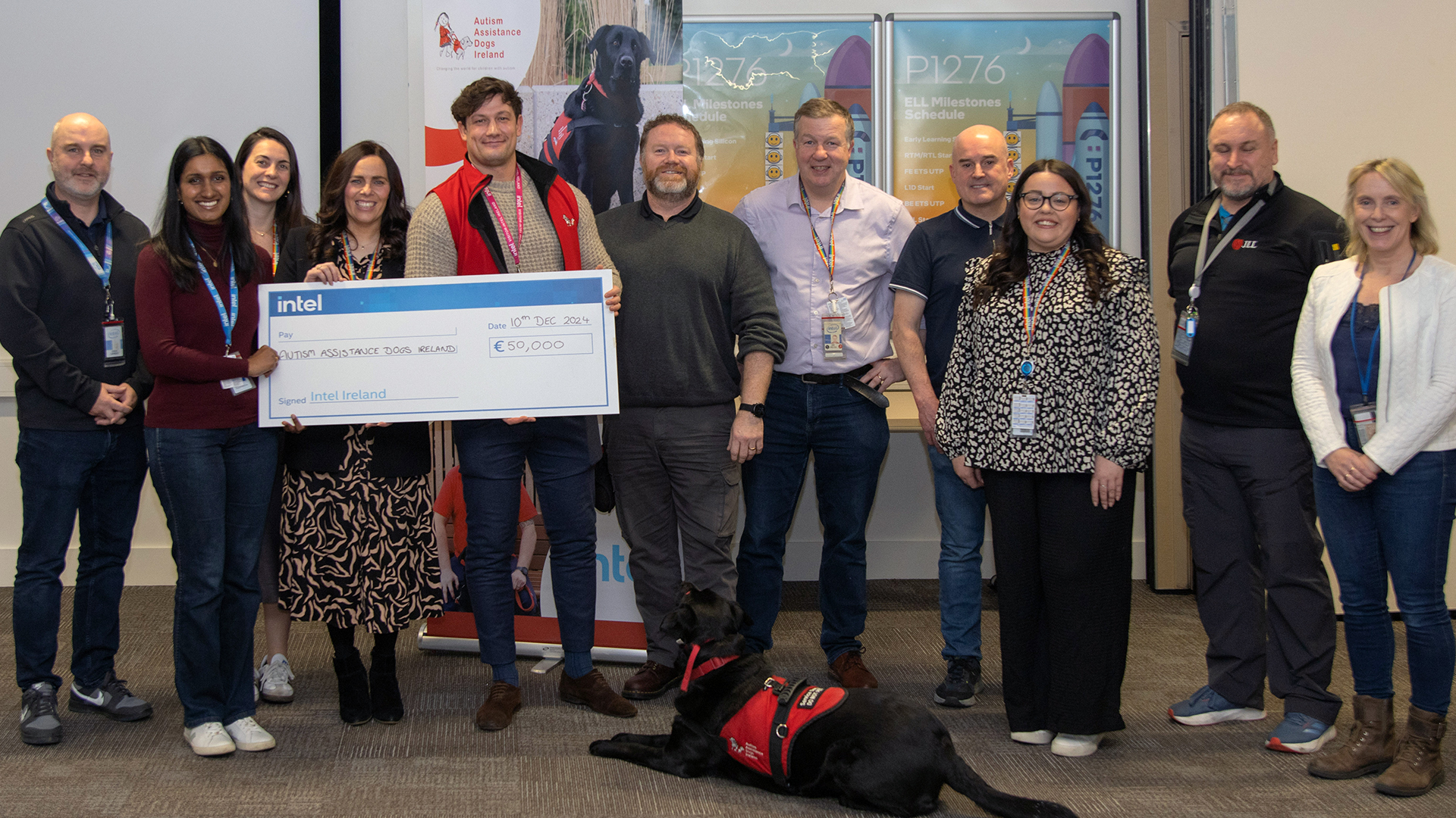 A group of people stand together indoors holding a large donation check for €5,000, made out to Autism Assistance Dogs Ireland. A service dog in a red harness sits on the floor in front of them. Various banners are visible in the background.