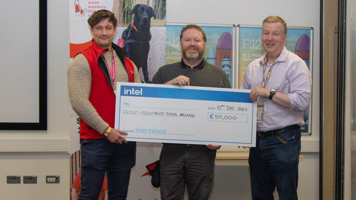 Three men stand indoors holding a large novelty check from Intel Ireland for €50,000, made out to Autism Assistance Dogs Ireland. A banner featuring a black dog and a rocket graphic is visible in the background.