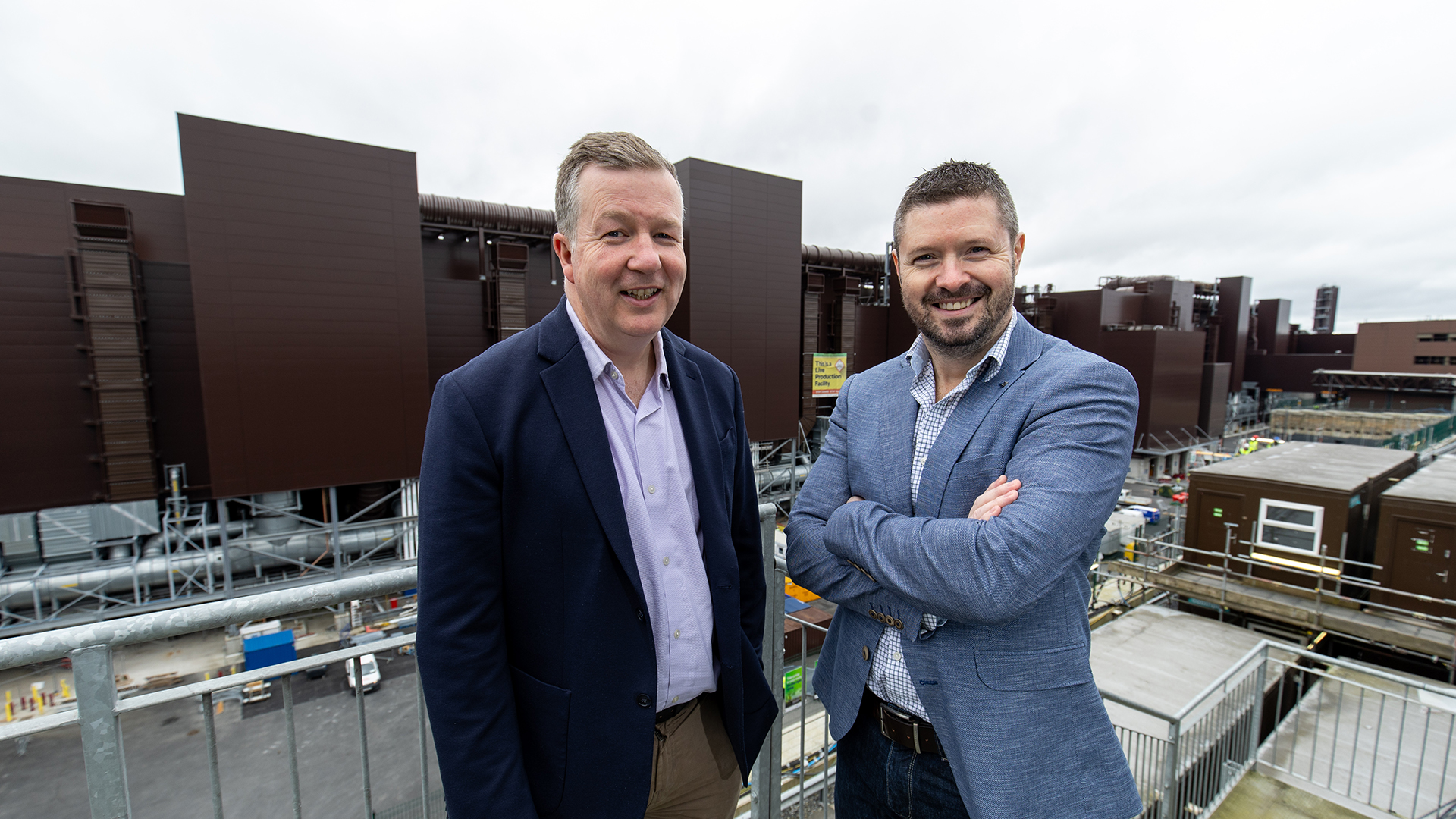 Two men in business attire stand on a rooftop with arms crossed, smiling at the camera. Behind them, large industrial buildings are visible under a cloudy sky.