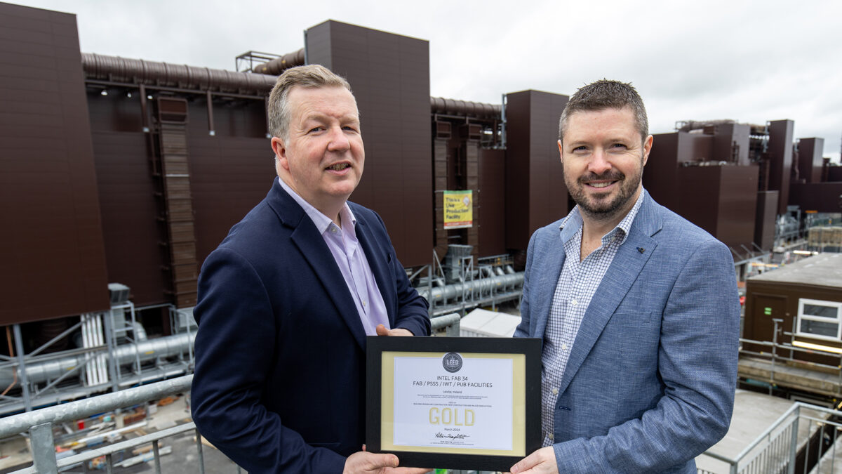 Two men in suits stand outside an industrial building, holding a framed certificate labeled Gold Free From Food Awards. The background shows large structures and overcast skies.