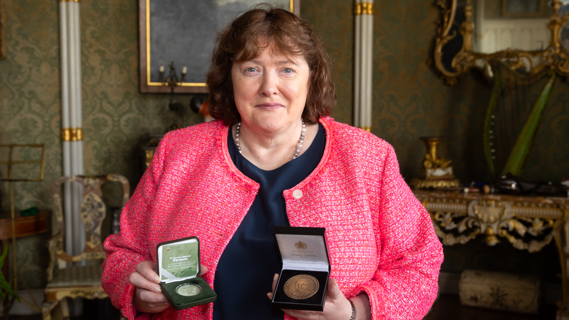 A woman in a pink jacket holds two open medal cases, displaying medals inside. The background features elegant, ornate decor with green wallpaper, a mirror, and antique furniture. She looks directly at the camera.