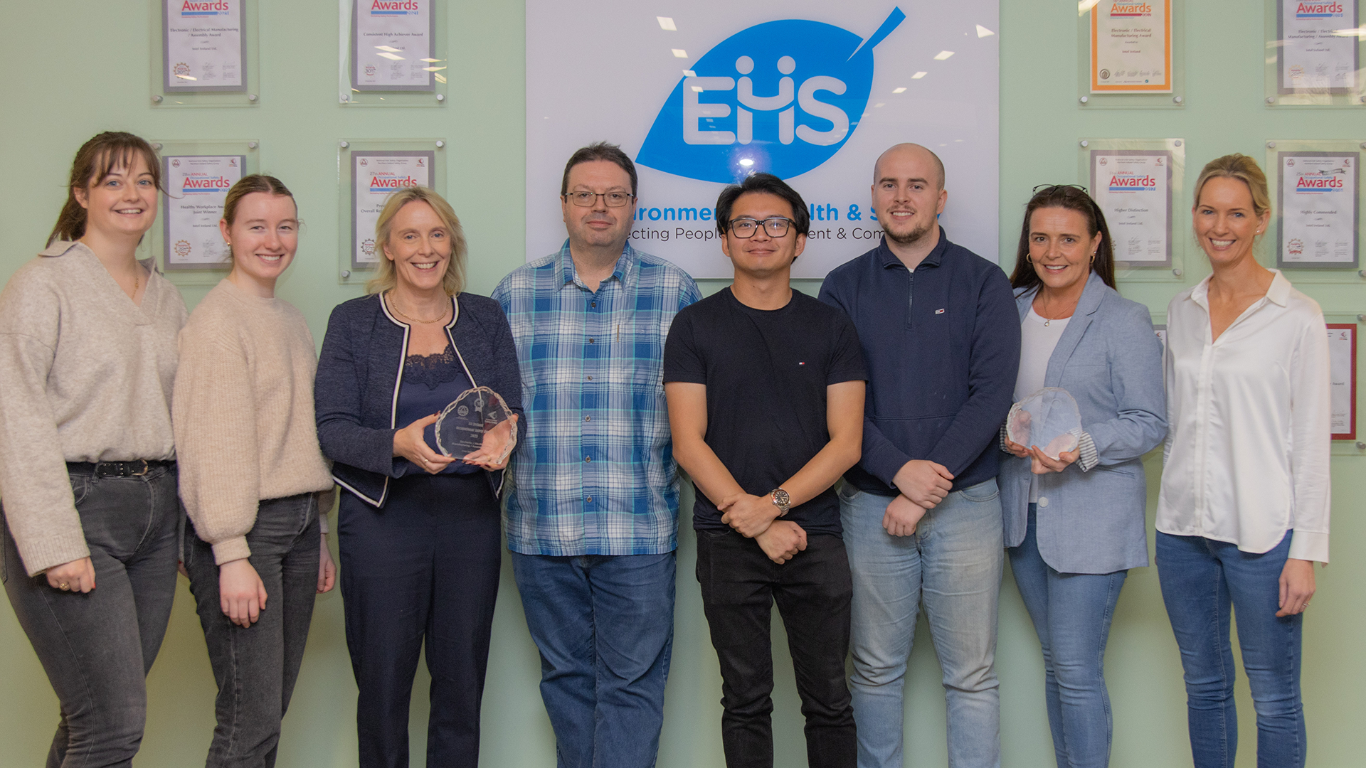 A group of nine people stands in front of a wall displaying award plaques. One individual holds the crystal transparent award. They are smiling and dressed casually. The backdrop features a logo reading EHS.