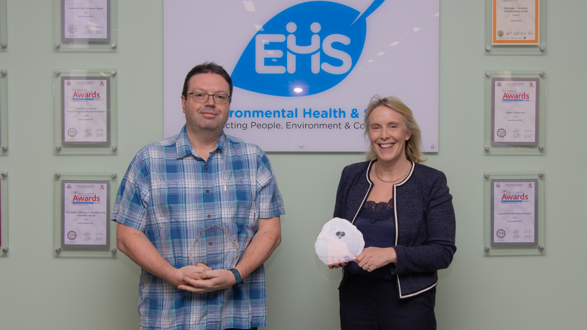 Two people holding awards stand in front of a wall featuring a logo and framed certificates. One person is wearing a blue plaid shirt; the other is in a dark blazer. A sign behind them reads Environmental Health & Safety.