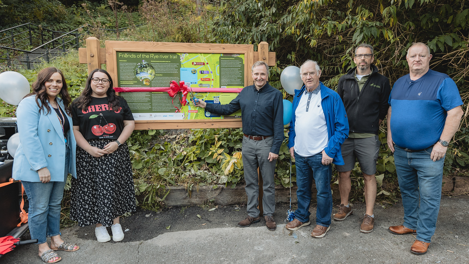 A group of six people stands outdoors in front of a large informational sign. The sign is decorated with a red ribbon and bow. Trees are in the background. The individuals are casually dressed and smiling at the camera.