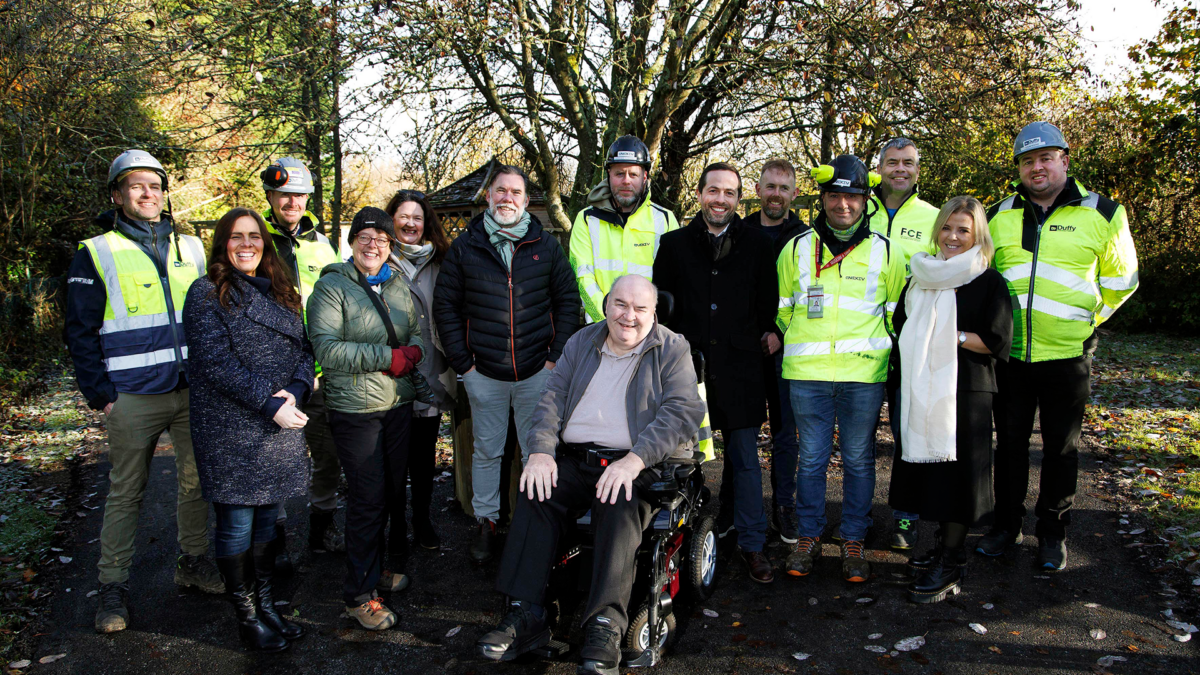 A group of people pose outdoors on a paved path surrounded by trees. Some are wearing high-visibility jackets and hard hats, while others are dressed in casual clothing. A man in a wheelchair is at the center. The setting is sunny and autumnal.