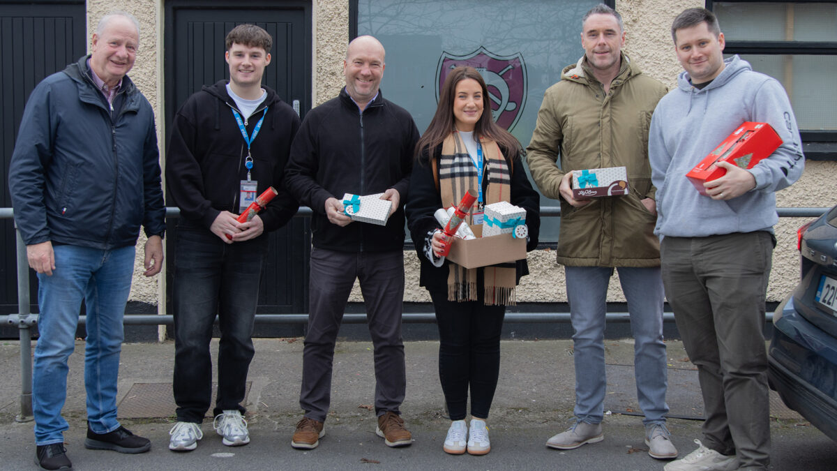 A group of six people standing outside a building, each holding small gift boxes and festive crackers. They are smiling and appear to be part of a holiday or charity event. The background features a textured wall and a window.