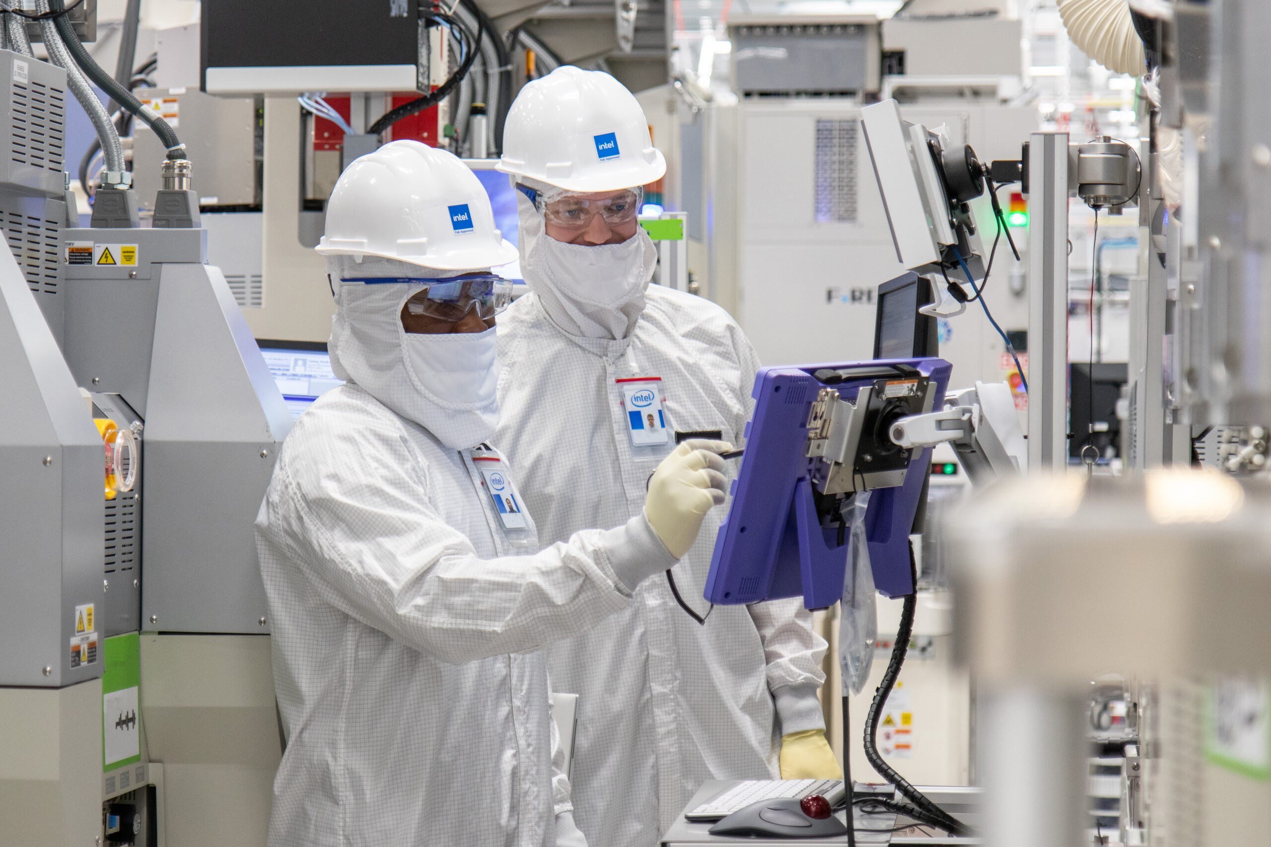 Two workers in cleanroom suits and helmets examine equipment in a high-tech manufacturing facility. One holds a tablet, while both are surrounded by complex machinery. Their attire includes protective masks and gloves.