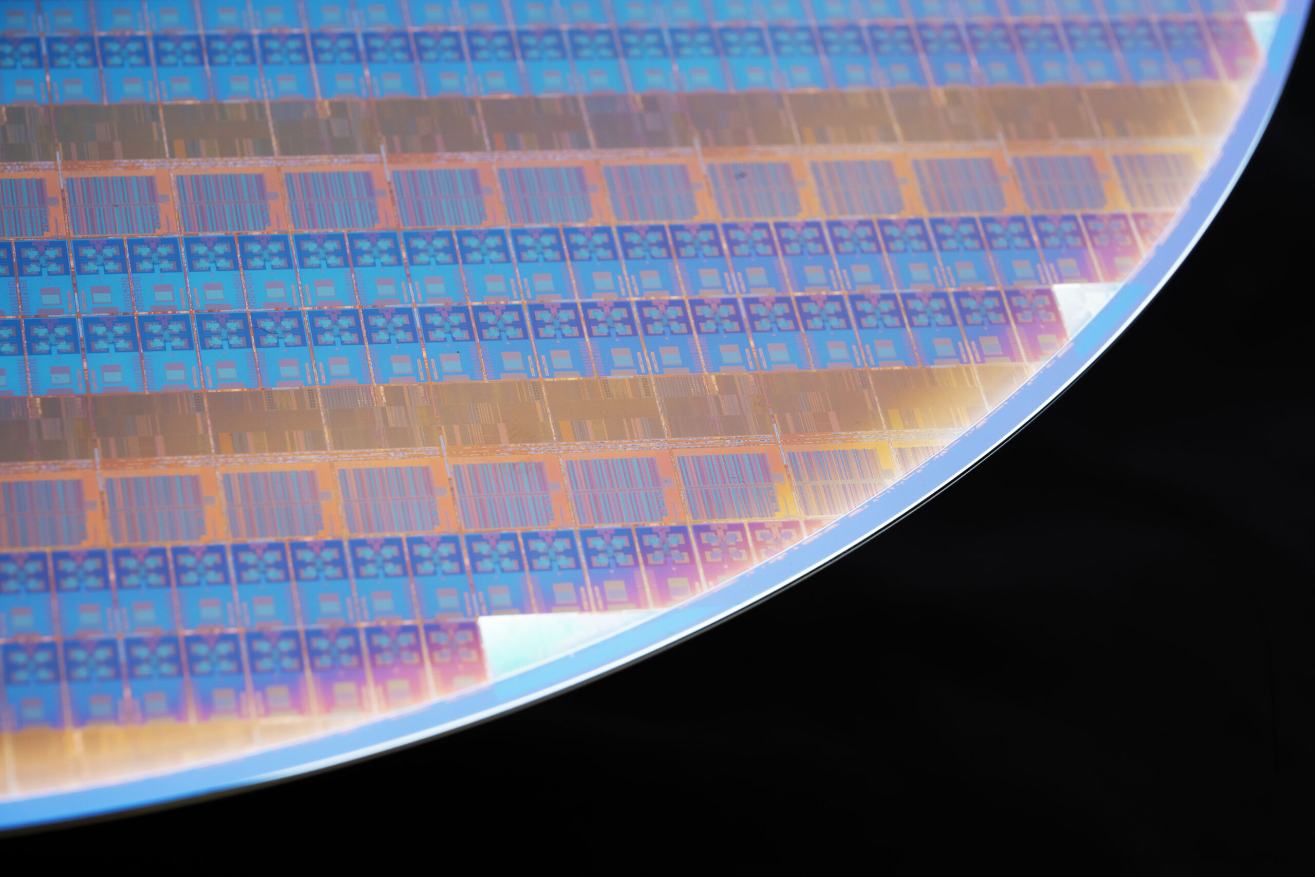 Close-up of a silicon wafer displaying intricate patterns of microchips in a grid layout. The wafer reflects various colors under the light, with a clear edge against a black background.