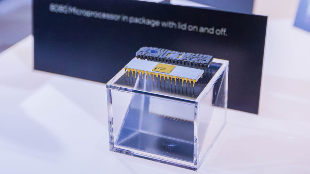 Close-up of a microprocessor displayed inside a clear, cubic case on a reflective surface. A blurred background features a sign with partial text visible, reading: processor in package with lid on and off.