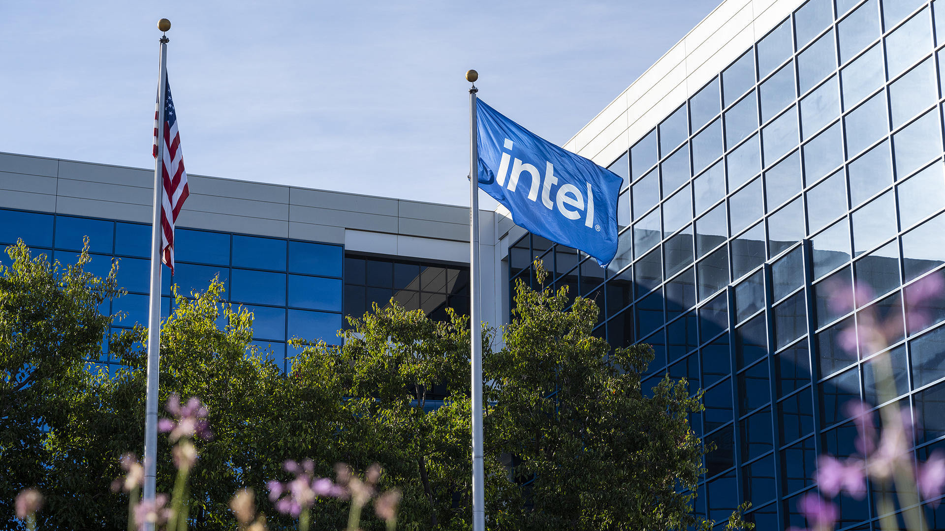 A blue flag with the Intel logo waves on a flagpole in front of a modern office building with reflective glass windows.