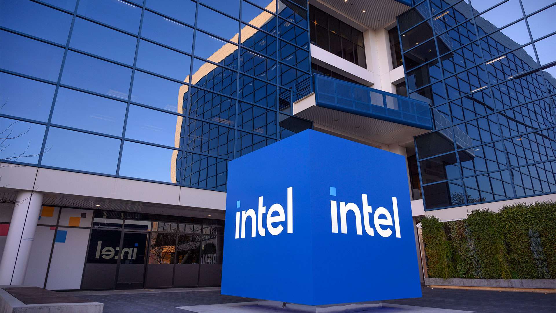 A large blue cube with the Intel logo sits in front of a modern office building with reflective glass windows.