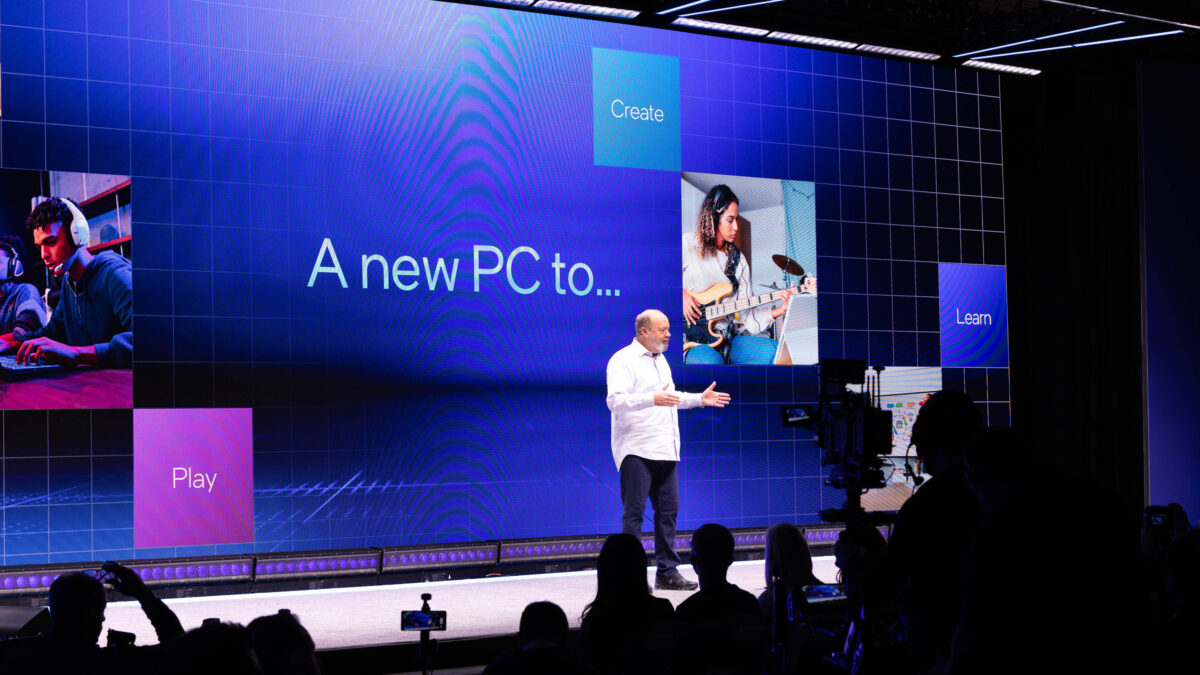 A speaker presents on stage in front of a large screen displaying A new PC to... with images and words: Create, Learn, Play. The audience is partially visible, along with cameras capturing the event.