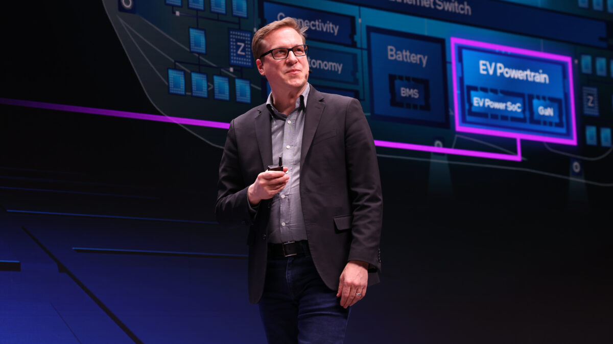 A man in glasses and a black blazer stands on stage holding a microphone. Behind him is a large screen displaying technical diagrams and terms like EV Powertrain and Battery. The setting appears to be a tech presentation or conference.