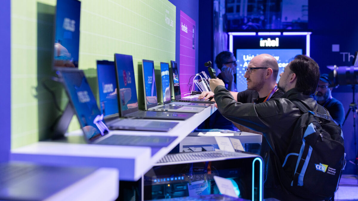 People are examining a row of laptops on display at an electronic exhibition. The background features bright lighting and signs, suggesting a tech-themed environment.