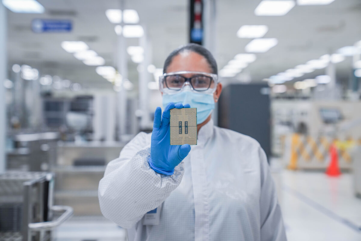 A person in protective clothing, including a face mask and gloves, holds a small electronic component in a lab or factory setting. The background shows industrial equipment, slightly blurred to emphasize the component.