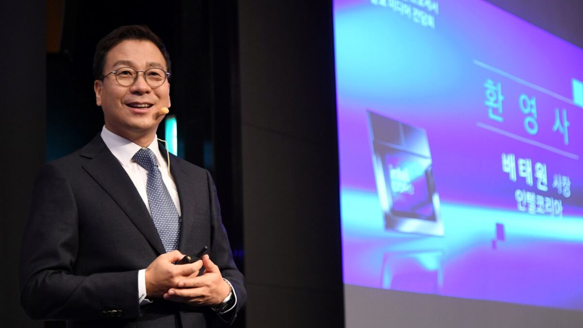 A man in a suit and tie stands smiling with a microphone on his ear. He holds a clicker and appears to be giving a presentation. A screen next to him displays Korean text and graphics.