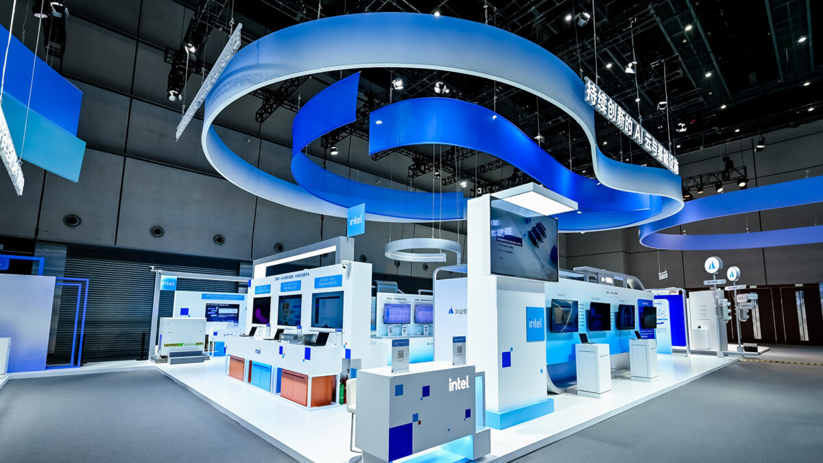 A modern exhibition booth with a sleek design features blue and white curved overhead structures. Various computer displays and informational panels with Intel branding are showcased throughout the space, surrounded by soft lighting.