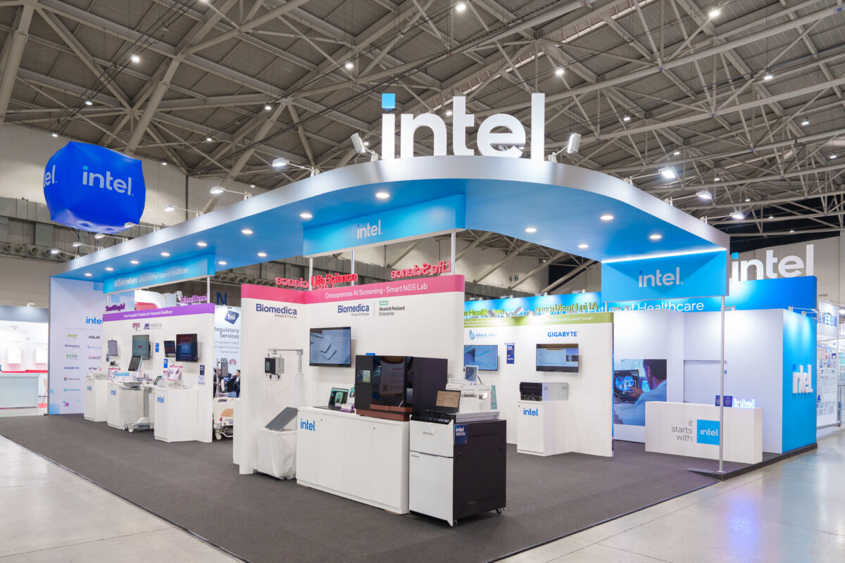 Wide-view of a tech exhibition stall branded with Intel logos. The booth features blue and white signage, product displays, digital screens, and informational panels. The open, modern design includes visitors interacting with exhibits.