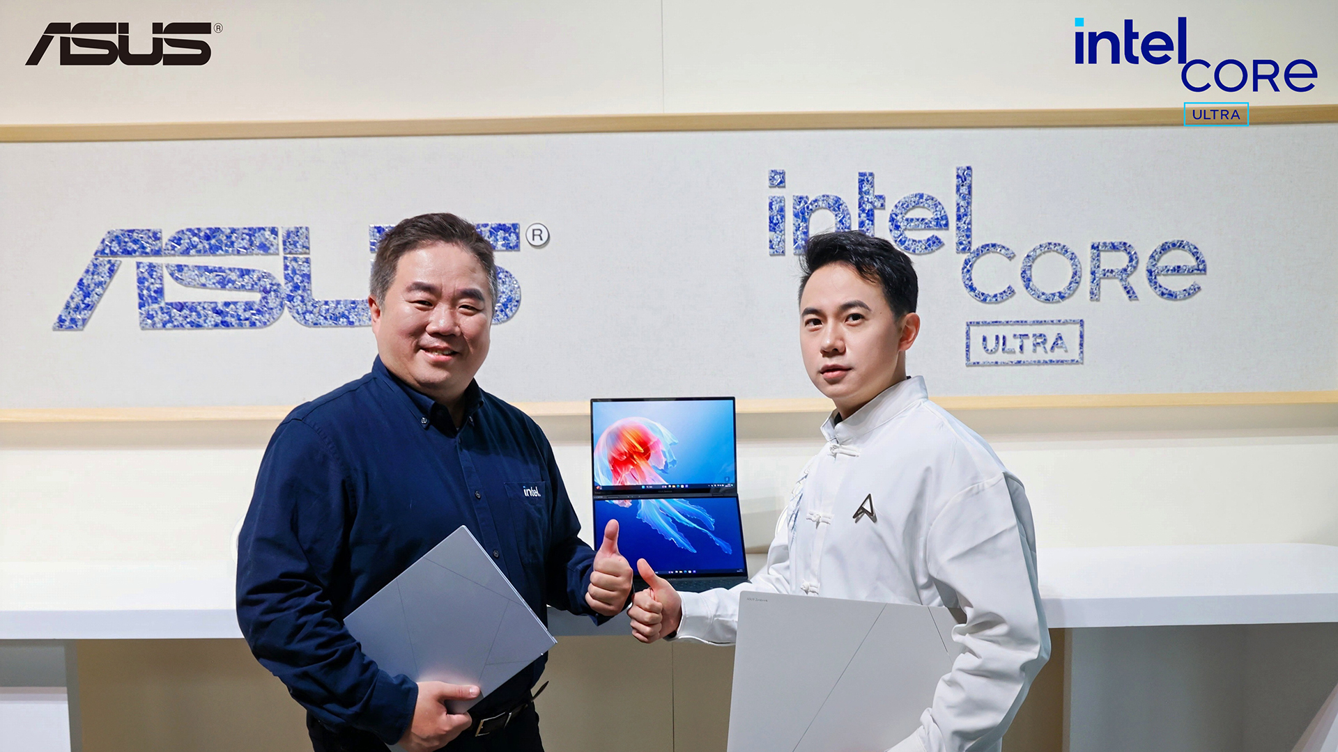 Two individuals holding laptops stand in front of a display featuring ASUS and Intel Core Ultra logos. One person gives a thumbs-up gesture. A laptop with a colorful screen is positioned between them. They are dressed in professional attire.