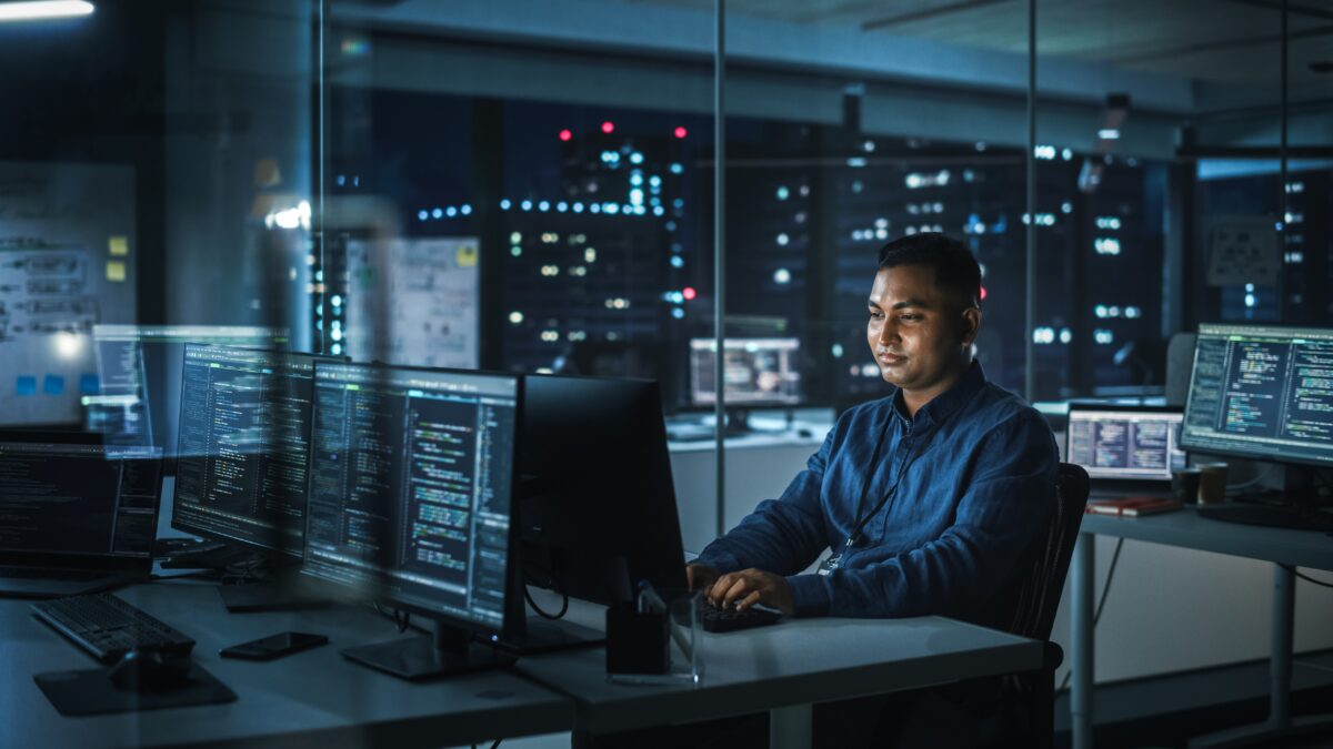 A person sits in a modern office at night, working on multiple computer screens displaying code. The room is dimly lit with reflections of city lights in the window, creating an atmosphere of focus and concentration.