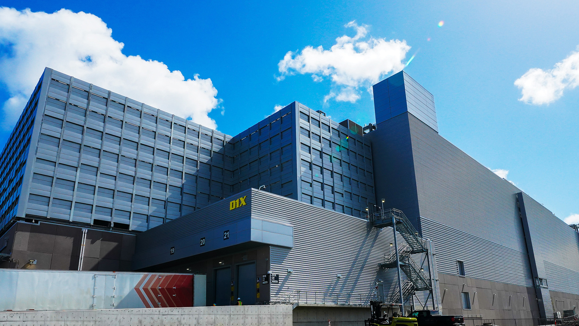 A modern industrial building with multiple floors is shown against a blue sky with scattered clouds. The structure features a combination of glass and metal surfaces, with a yellow sign displaying DIX on one section. A stairway is visible on the side.